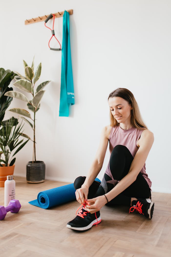 Full body young female athlete in sportswear tying shoelaces while sitting on floor near sport equipment and potted plants and looking down with smile